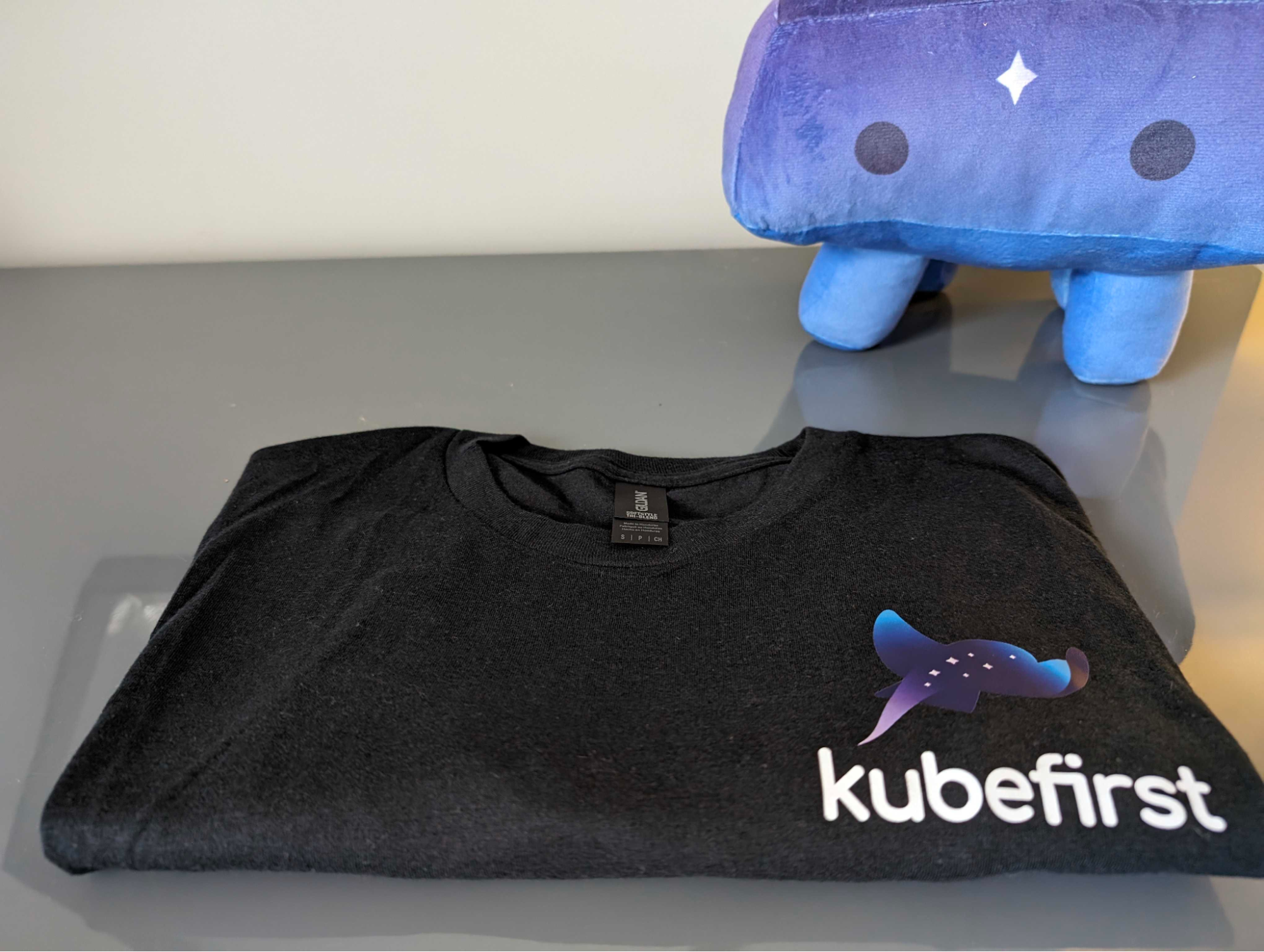a black folded t-shirt with the kubefirst logo with the K-Ray plushy in the back.